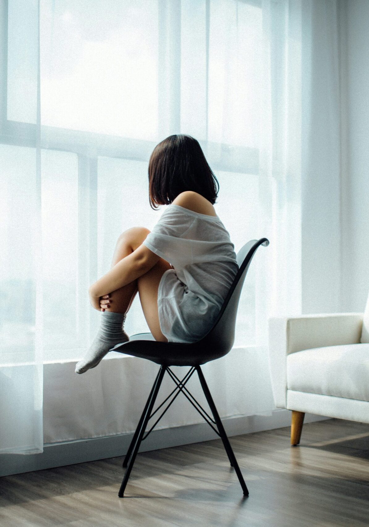 Woman sitting on black chair in front of glass panel window with white curtains.